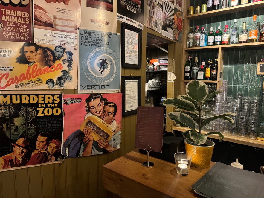 Wall at Elly Bar decorated with classic film posters including Casablanca and Vertigo, beside a bar counter with glassware and bottles.