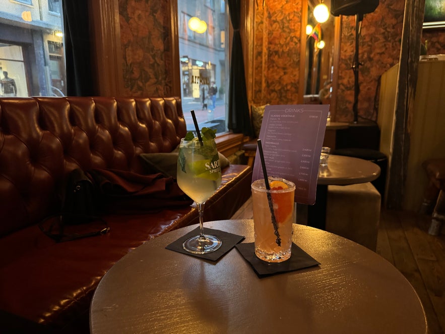 Two colorful cocktails on a round table at Elly Bar, set against a red leather banquette and soft evening lighting.