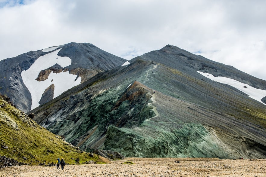 Hikers climbing Blahnukur Mountain in Landmannalaugar, Iceland, with colorful rhyolite slopes and snow patches.