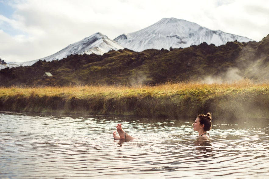 Traveler relaxing in a natural hot spring at Landmannalaugar, Iceland, with snowy volcanic mountains in the background.