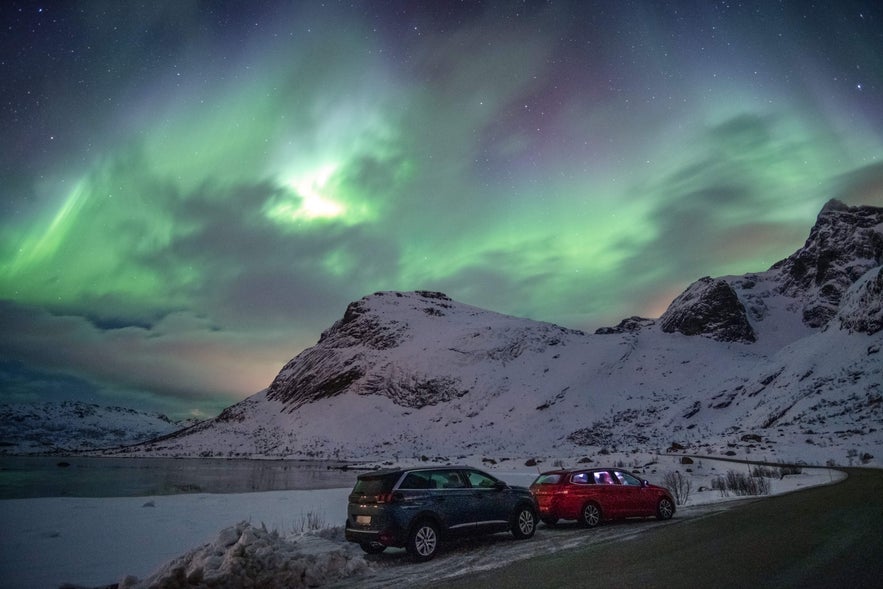 Cars parked on a snowy roadside under the northern lights in Iceland.