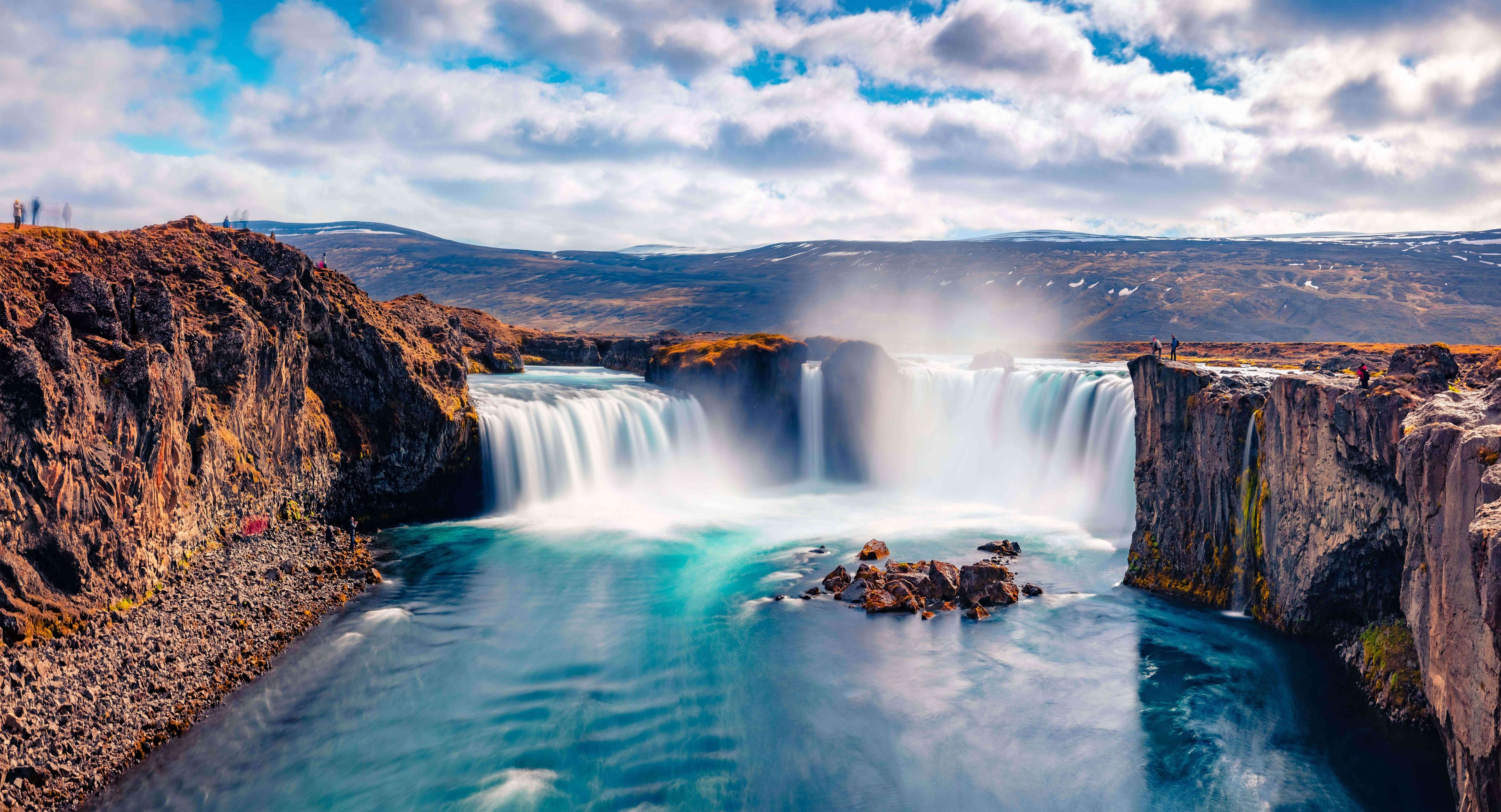 Godafoss-vandfaldet i Nordisland, der falder ned i turkisblåt vand, med brede vandmasser og klipper under en delvist overskyet himmel.