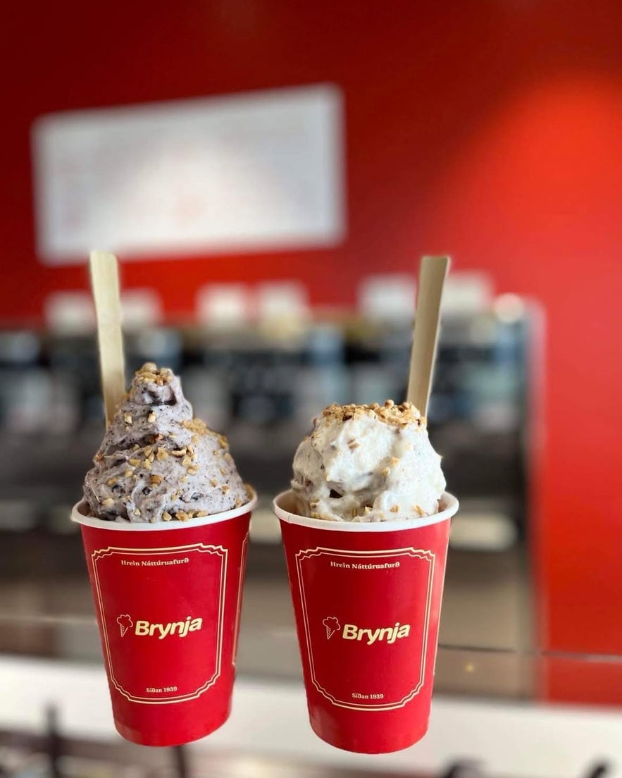 Two cups of Brynja ice cream with toppings and wooden spoons on a counter, set against a red shop interior.