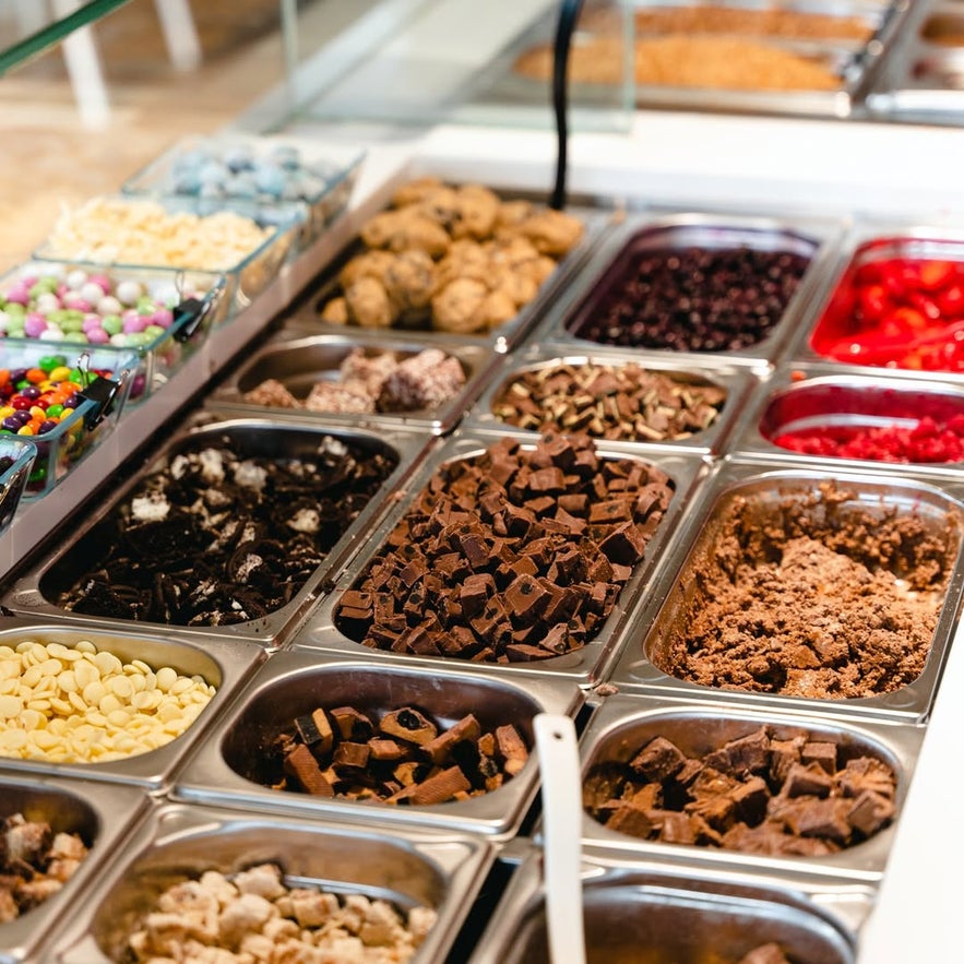 Brynja toppings bar with cookies, candies, marshmallows, and chocolate pieces in metal containers for self-serve delight.