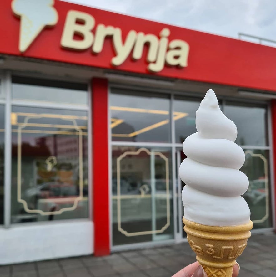 Brynja soft-serve cone in front of the shop’s red-and-white facade with bold cream signage and decorative gold trim.