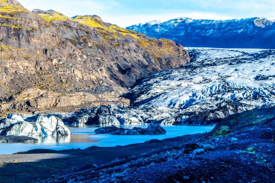 Näkymä Sólheimajökull-jäätikölle ja sen laguunille Etelä-Islannissa; jäämuodostelmia ja vulkaanisia rinteitä kirkkaassa auringonvalossa.
