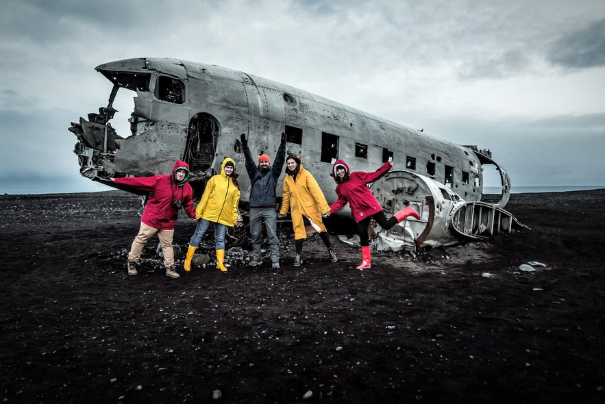 Group of travelers in colorful jackets posing beside the DC3 Plane Wreck on Solheimasandur Beach in Iceland.
