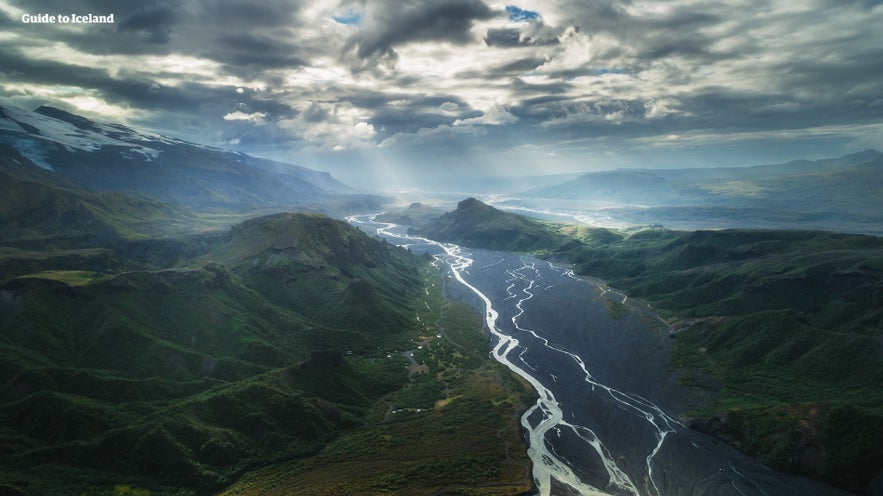 A river flows through Thorsmork Valley, surrounded by lush greenery and under a blanket of cloudy skies.