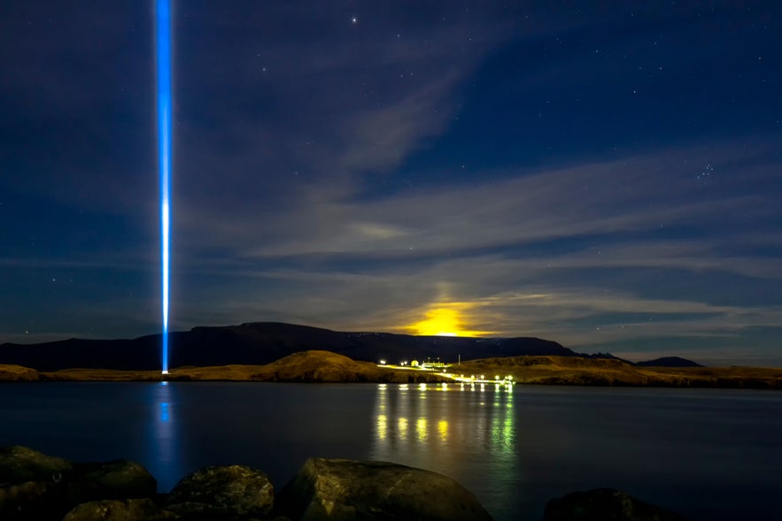 Imagine Peace Tower in Reykjavik beams blue light into starry sky, with moonrise over water and distant mountains. Imagine Peace Tower in Reykjavik beams blue light into starry sky, with moonrise over water and distant mountains.