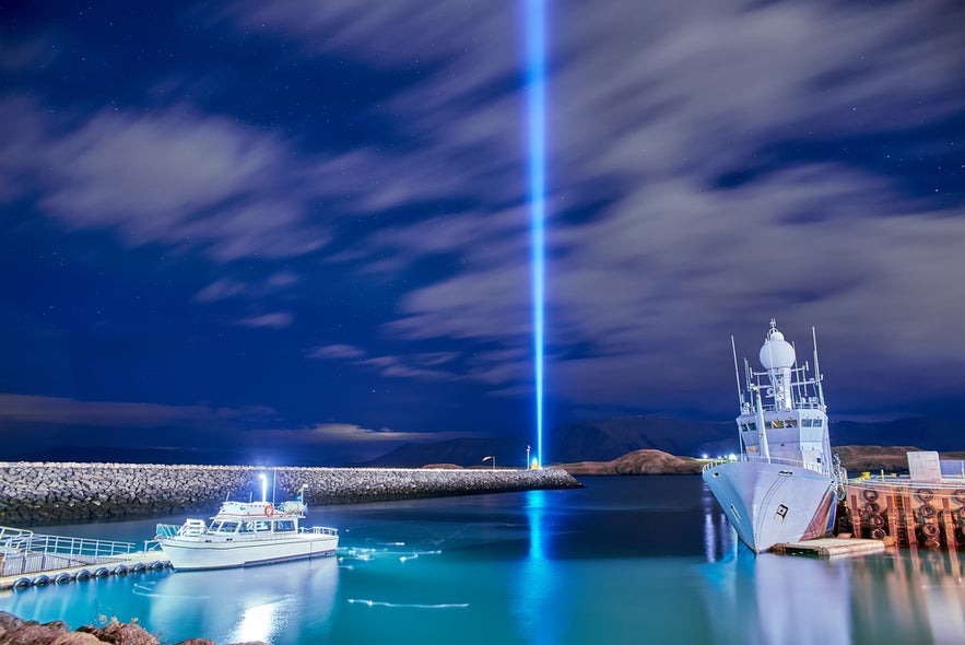 Imagine Peace Tower beams blue light into Reykjavik’s night sky, with docked boats and calm harbor reflecting the glow Imagine Peace Tower beams blue light into Reykjavik’s night sky, with docked boats and calm harbor reflecting the glow
