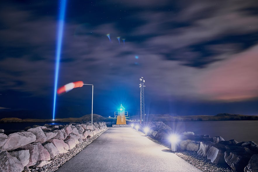 Nighttime pier in Reykjavik with windsock and beacon, as Imagine Peace Tower beams blue light into starry sky. Nighttime pier in Reykjavik with windsock and beacon, as Imagine Peace Tower beams blue light into starry sky.