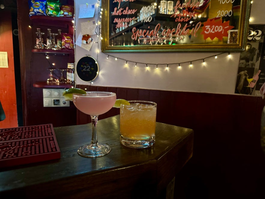 Two cocktails on the bar counter at 22 Bar, one pink with lime garnish and one amber-colored, with string lights and mirrors in the background.