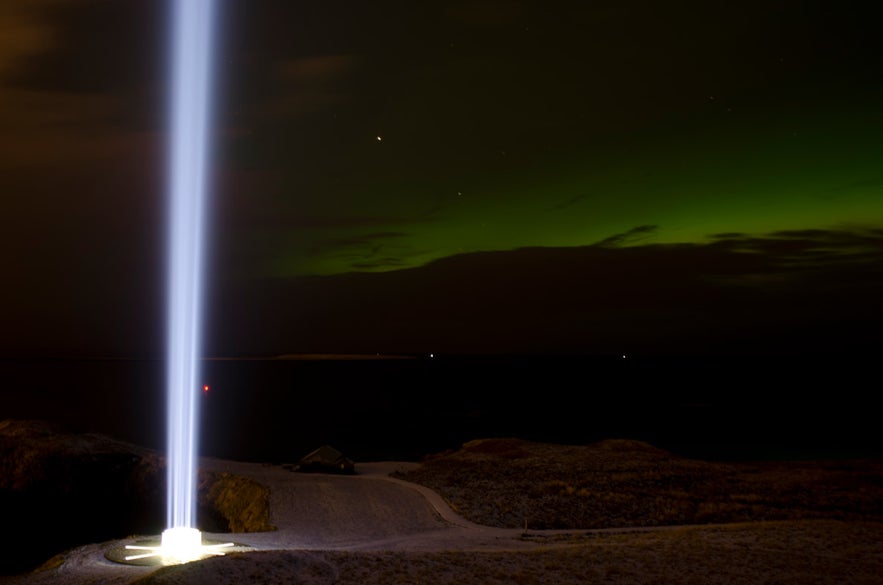 Imagine Peace Tower’s beam shines over Videy Island at night with the green glow of the northern lights in the sky.