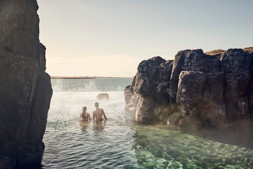 Two people relax in the warm infinity pool at Sky Lagoon in Reykjavik, surrounded by rocky cliffs and ocean views.