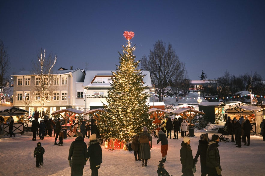 People explore the Christmas Village in Hafnarfjordur, with a large lit tree, market stalls, and snowy festive decorations.
