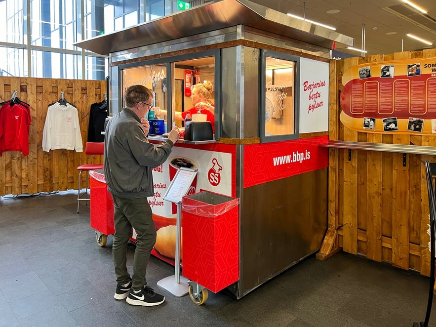 Traveler buying an Icelandic hot dog at Baejarins Beztu Pylsur stand inside Keflavik Airport during a layover in Iceland.