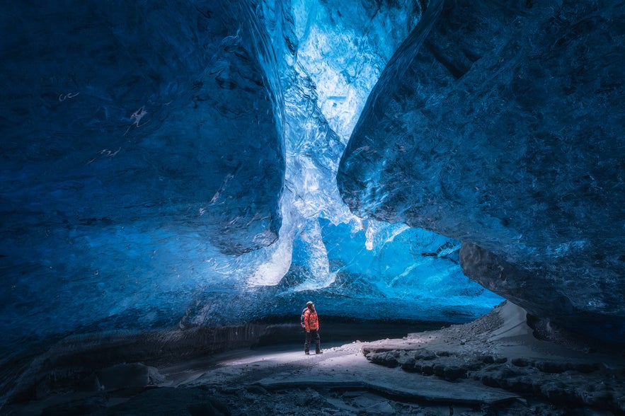 A person in a red jacket stands inside Iceland’s Crystal Ice Cave, surrounded by deep blue glacial walls and translucent ice formations glowing in natural light.