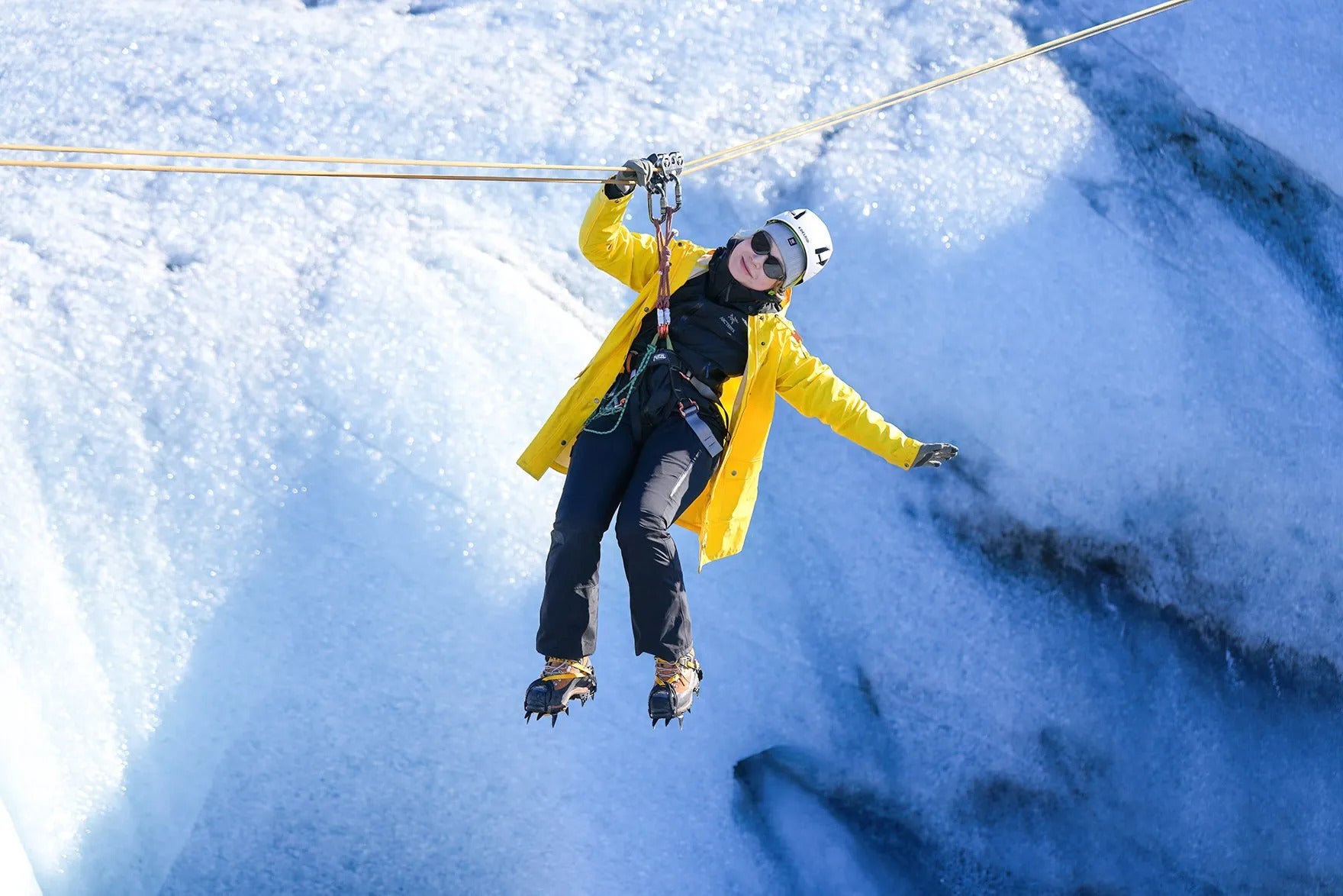 Woman in a yellow jacket glides on a rope over the icy blue formations of Solheimajokull Glacier.