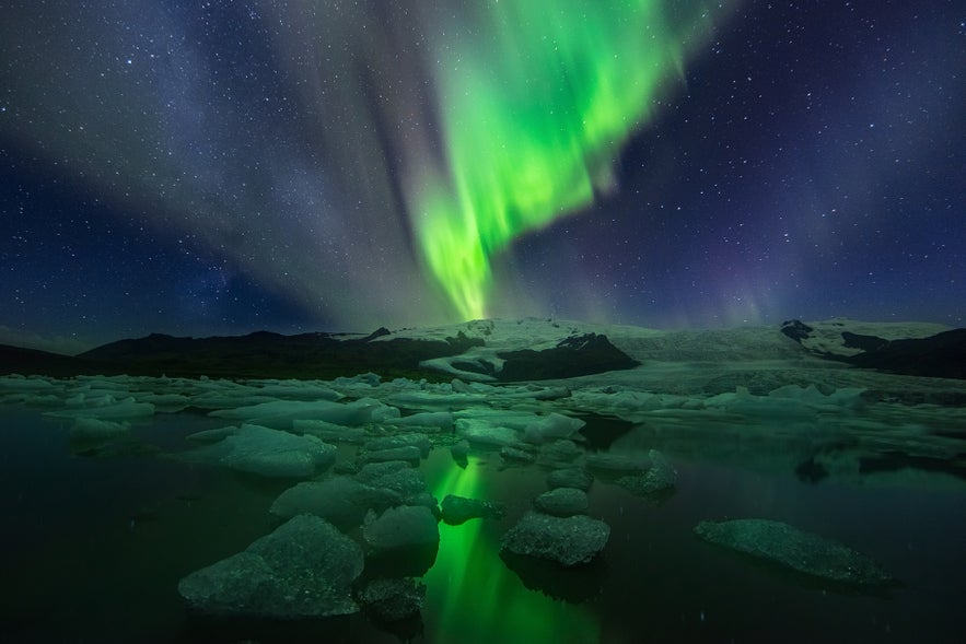 A vivid display of the northern lights glows green over Fjallsarlon Glacier Lagoon in southeast Iceland, reflecting across floating icebergs with Vatnajokull Glacier and a star-filled night sky in the background.