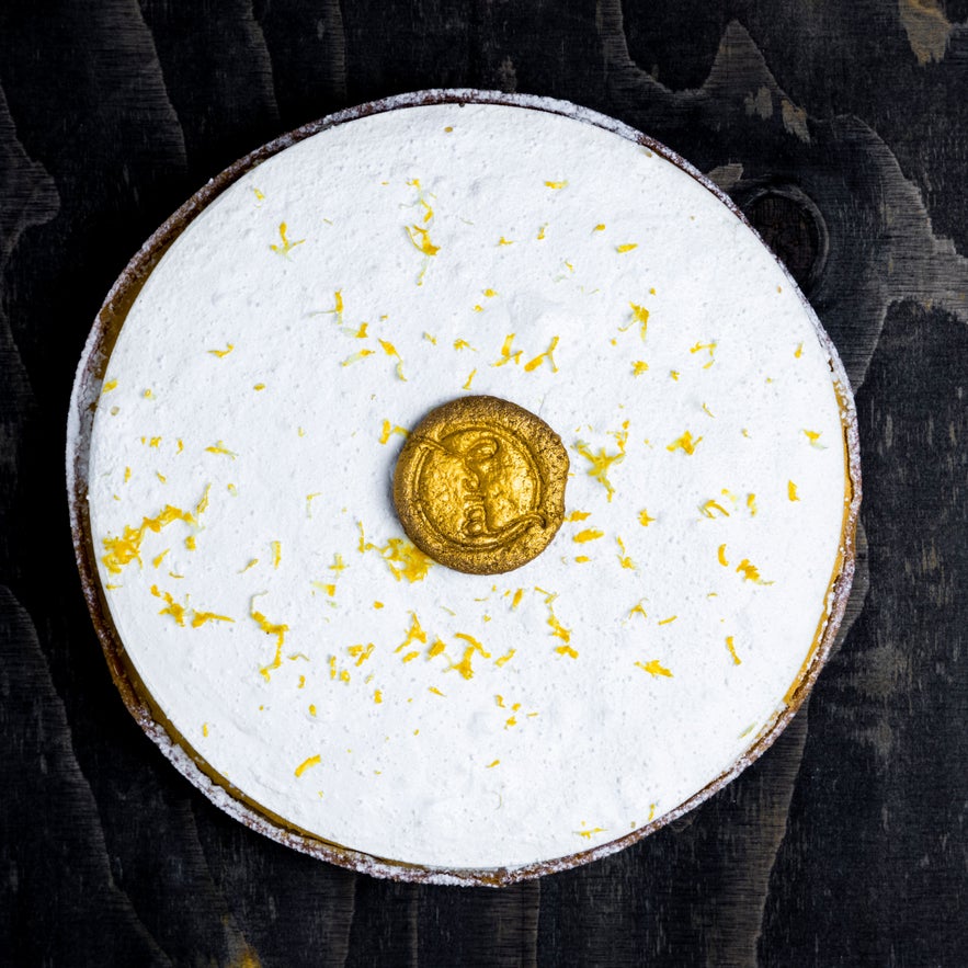 ChatGPT said:  Top view of a white frosted cake with yellow zest and a gold Sweet Aurora seal on a dark wooden surface.