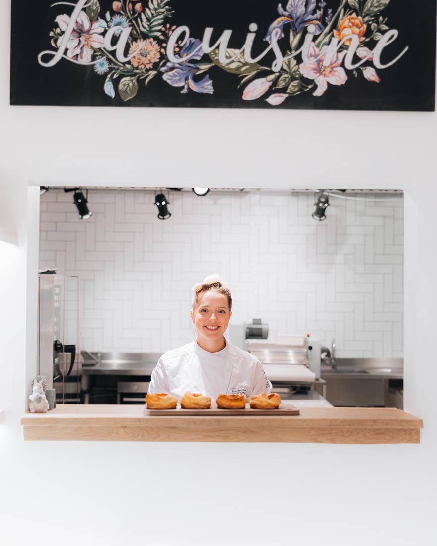Chef at Sweet Aurora stands behind a counter with five pastries, beneath a 'La cuisine' sign in a modern kitchen.