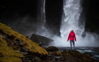 Base of Kvernufoss Waterfall in Iceland, with water cascading down a moss-covered cliff into a shallow pool surrounded by lush greenery.