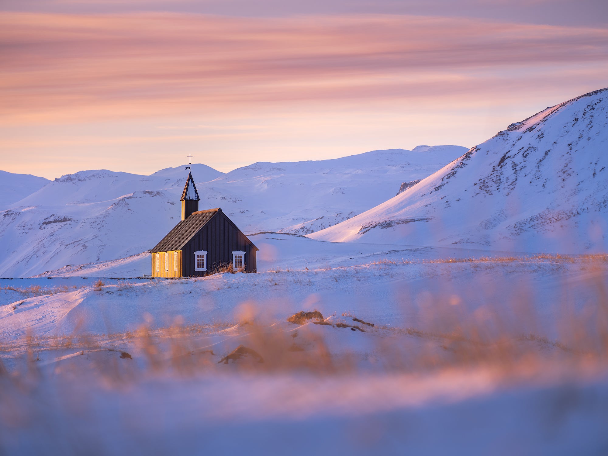 Budakirkja Black Church surrounded by snow and mountains under a colorful winter sunset sky.