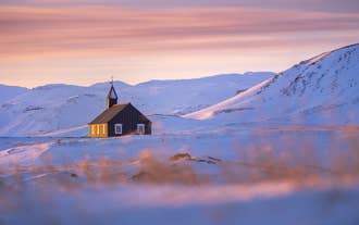 Budakirkja Black Church surrounded by snow and mountains under a colorful winter sunset sky.