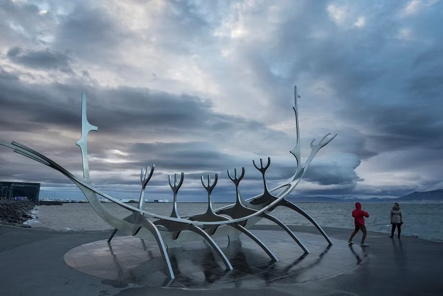 A person is posing for a photo behind the Sun Voyager Sculpture in Reykjavik while the other person takes a photo. 