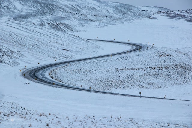 A winding road is surrounded by snowy fields on a winter day in Iceland. 