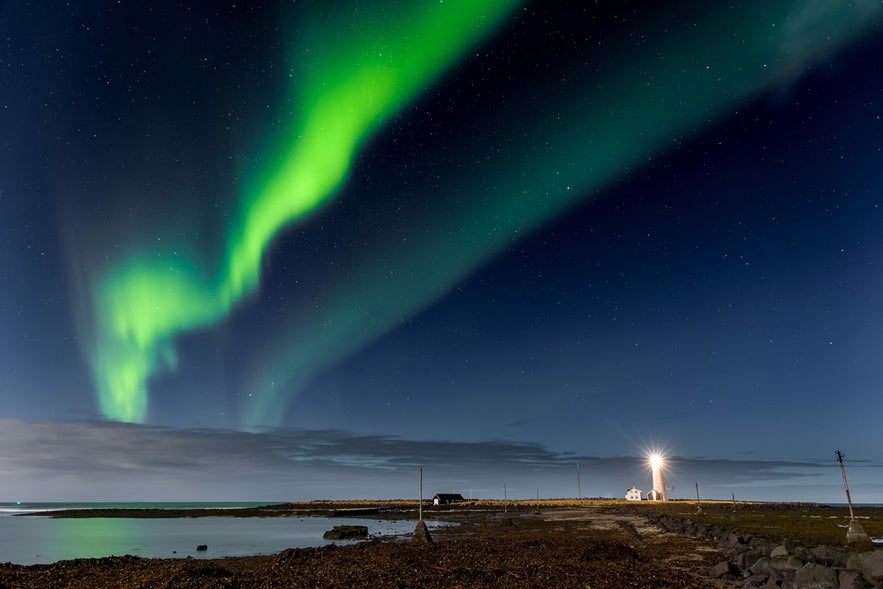Northern lights glowing above Grotta Lighthouse near Reykjavik in April with reflections over the coast.
