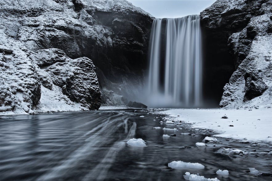 Snow blankets the area around Skogafoss Waterfall with icy mist rising from the falls.