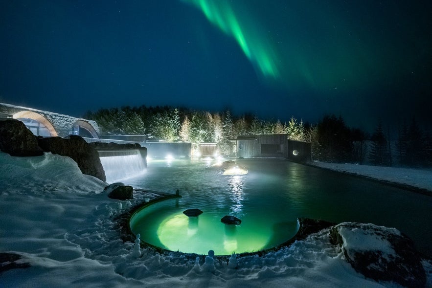 Northern lights above the geothermal pools of Laugarás Lagoon in Iceland, surrounded by snow and winter forest