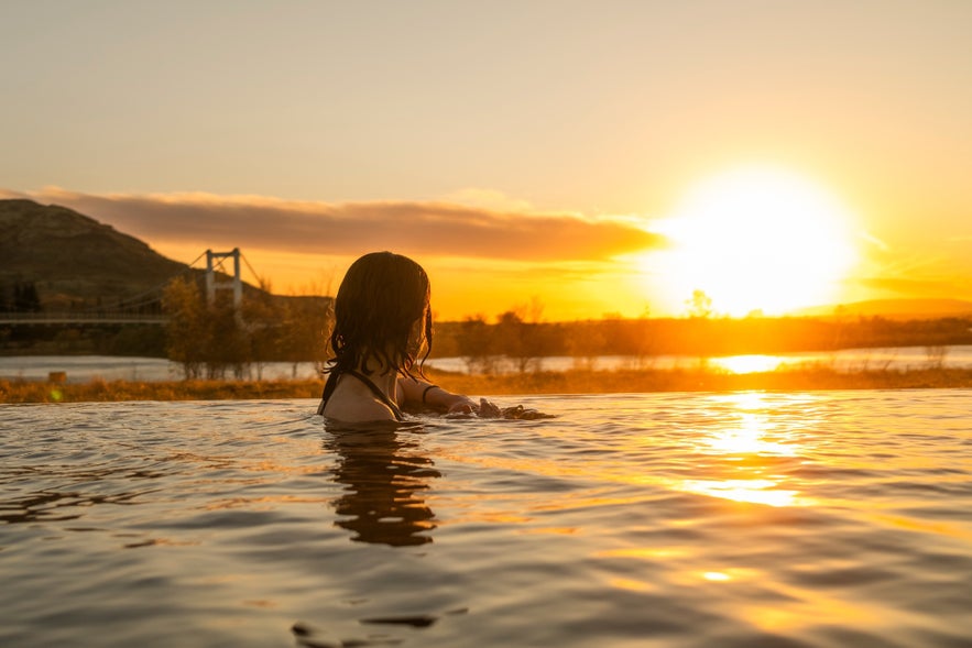 G&aelig;st, der slapper af i det geotermiske vand i Laugar&aacute;s Lagoon ved solnedgang med bro og bjerge i baggrunden