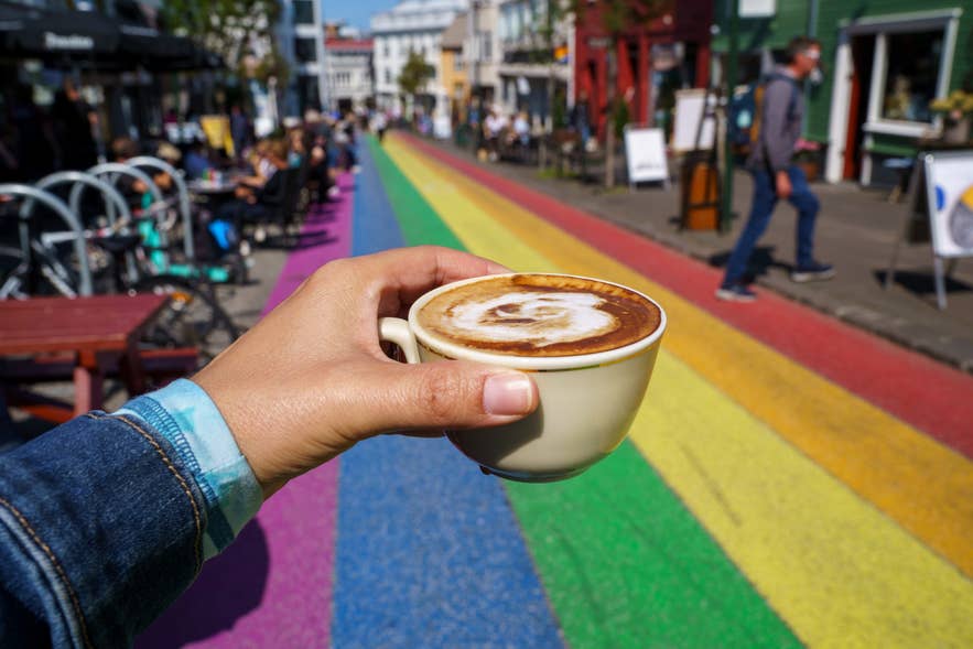 A hand holding a cup of coffee on Reykjavik’s rainbow-painted street with colorful buildings and outdoor cafes in the background.