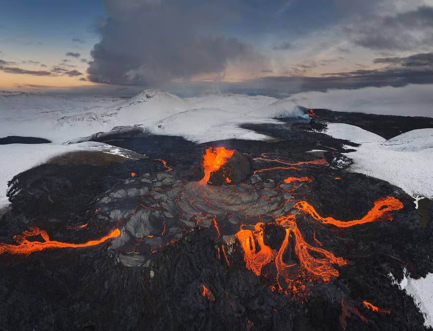 Aerial view of a volcanic eruption in Iceland, with glowing lava flows cutting through dark rock and snow-covered mountains in the background.