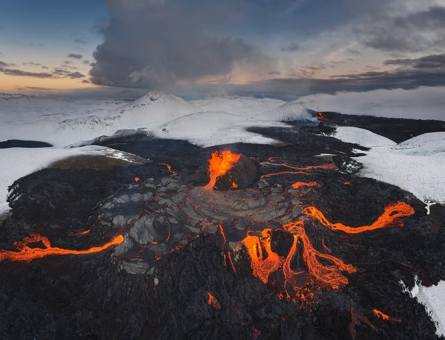 Aerial view of a volcanic eruption in Iceland, with glowing lava flows cutting through dark rock and snow-covered mountains in the background.