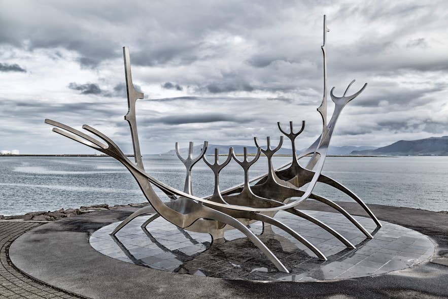 The Sun Voyager sculpture in Reykjavik, Iceland, standing beside the waterfront with mountains and cloudy skies in the background.