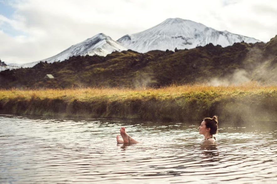 Person relaxing in a natural hot spring at Landmannalaugar in Iceland, with steam rising and snow-covered mountains in the background.