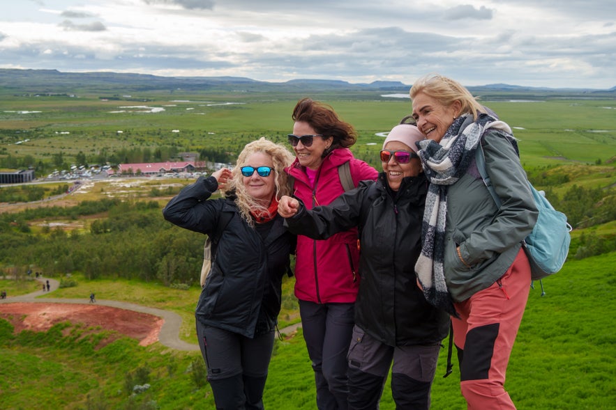 Group of four women enjoying a hike at Thingvellir National Park in Iceland, smiling with a scenic green valley and mountains in the background.