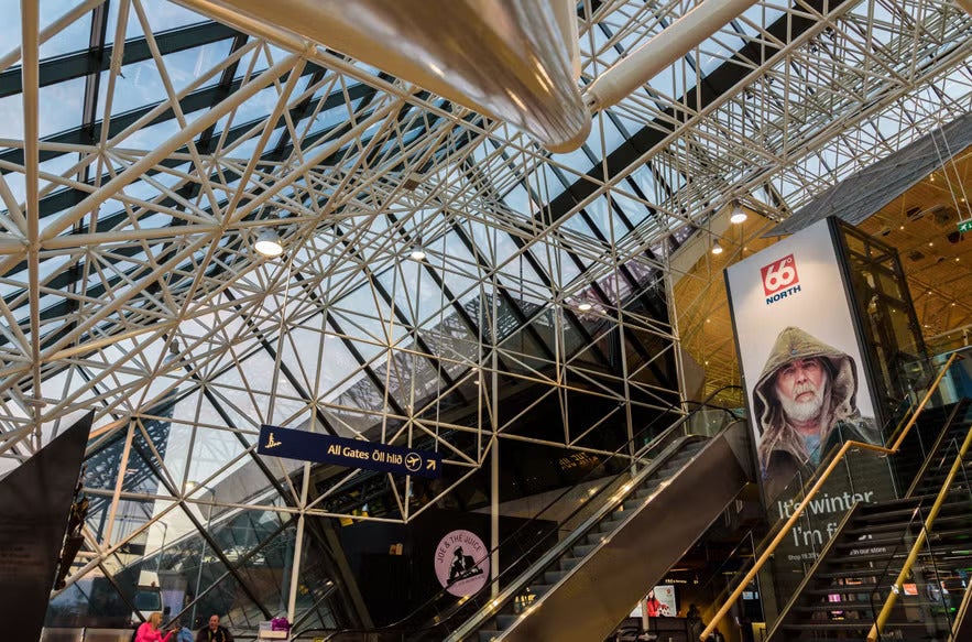 Interior of Keflavik International Airport in Iceland, showing modern glass architecture, escalators, and a 66°North advertisement.