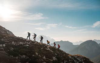 A group hikes uphill along a rocky slope with trekking poles against a bright sky and distant Icelandic mountain peaks.