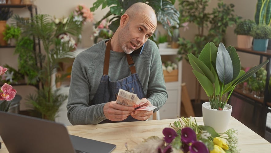 A man counting Icelandic krona in a flower shop while on the phone, illustrating small business work in Iceland. 
