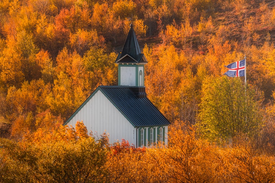 Rear view of Thingvallakirkja Church framed by autumn foliage, with an Icelandic flag visible on the right.