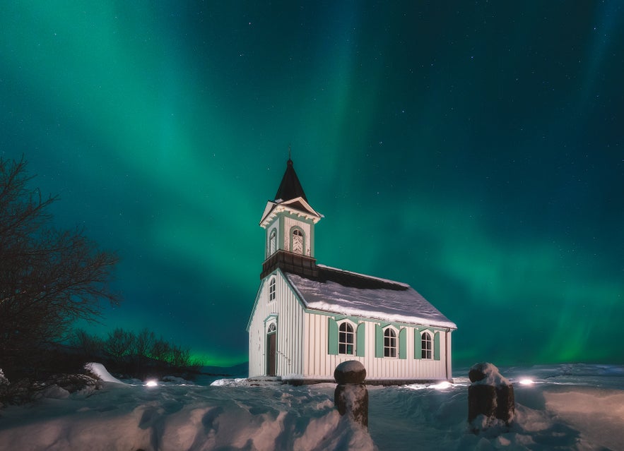 Thingvallakirkja, a small white wooden church with green accents, stands in deep snow beneath bright green northern lights in Thingvellir National Park.