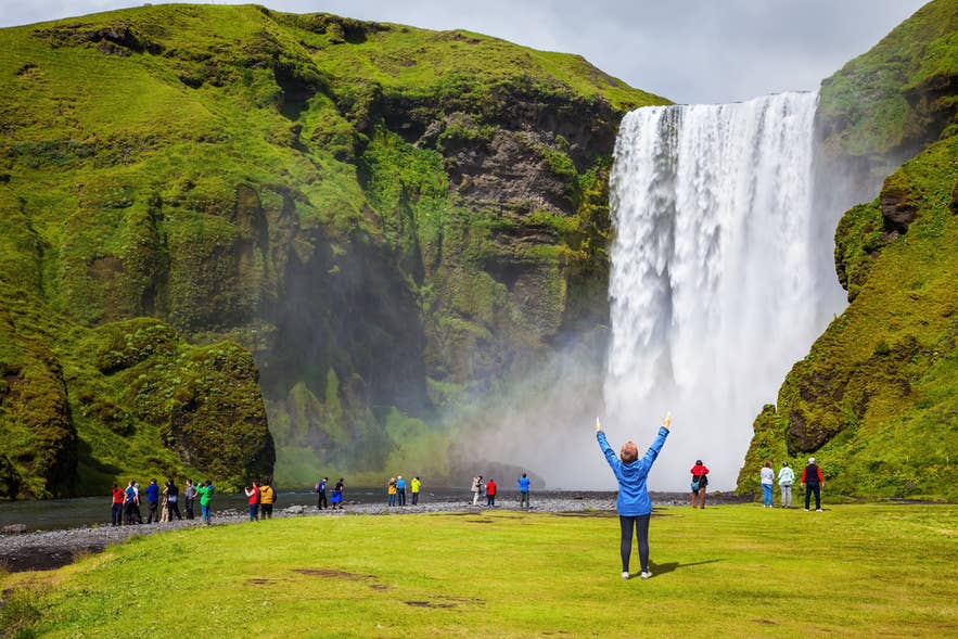 A tour group admiring and taking photos near Iceland waterfalls with green cliffs and clear summer sky.