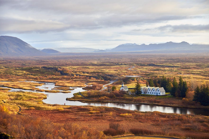 A high-angle view of Thingvellir National Park in autumn, showing the Thingvallakirkja Church, the Thingvallabaer Manor, and Thingvallavatn Lake surrounded by colorful foliage.
