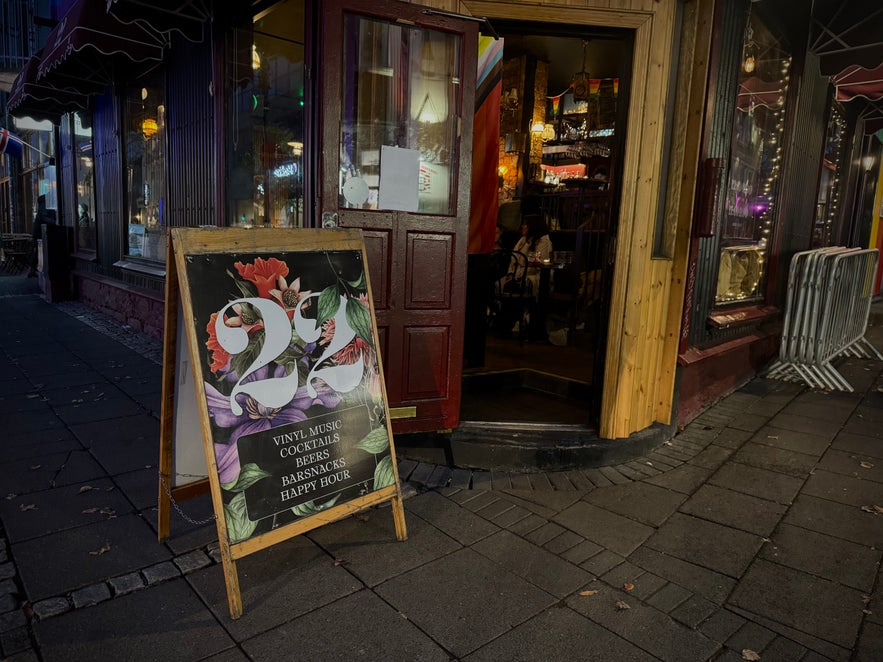 Entrance to 22 Bar on Laugavegur in Reykjavik, with the door open to a warmly lit interior and a sidewalk sign advertising vinyl music, cocktails, beers, bar snacks, and happy hour.