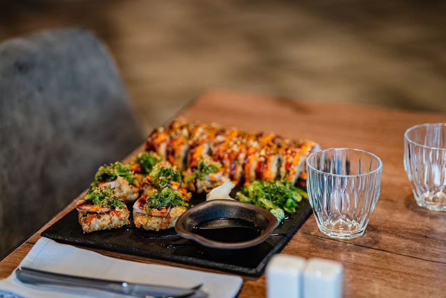 A sushi-style sharing platter served on a black slate at Glóð Restaurant, set on a wooden table with seaweed salad, dipping sauce, and two empty glasses beside it.