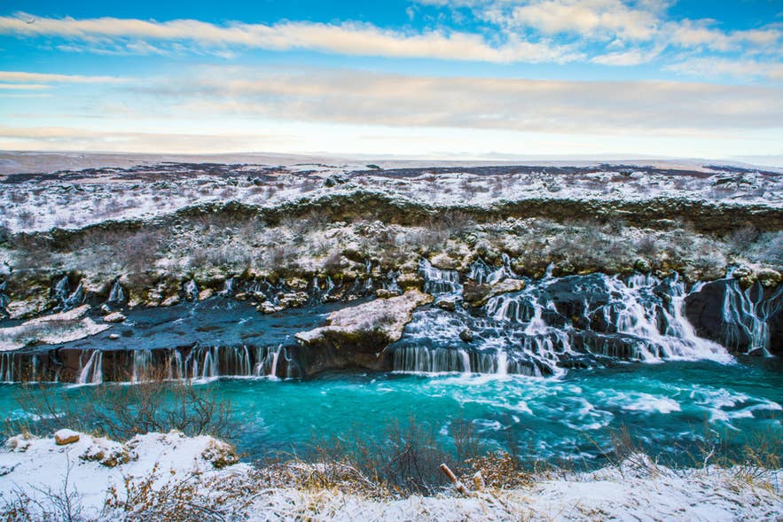 La cascata Hraunfossar scorre dalla roccia lavica nel fiume Hvita dalle acque turchesi, incorniciata dalla neve e dalla tenue luce invernale.